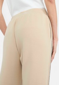 Pantalons de jogging beige avec une taille élastique, des poches latérales et une bande brillante le long des coutures extérieures pour un détail supplémentaire.