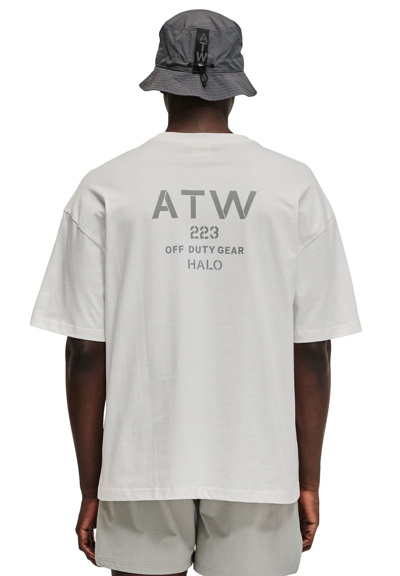 Weißes, übergroßes T-Shirt mit grauem Text, das "ATW 223 OFF DUTY GEAR HALO" auf dem Rücken trägt. Kombiniert mit einem grauen Bucket Hat.