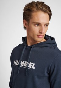 Młody mężczyzna z krótkimi, jasnobrązowymi włosami, ubrany w granatową bluzę z kapturem z logo "HUMMEL", patrzący zamyślonym wzrokiem w prawo na białym tle.