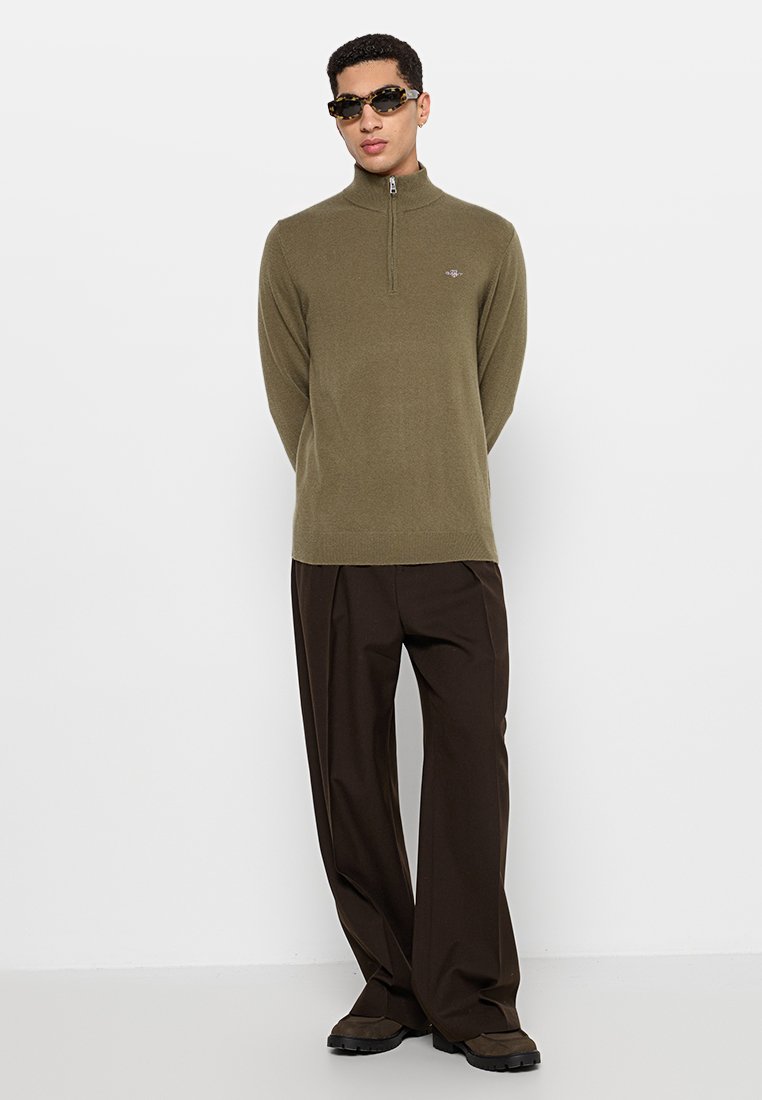 Mężczyzna w oliwkowym swetrze na zamek, ciemnobrązowych szerokich spodniach, brązowych butach i okularach przeciwsłonecznych w muszelkowym wzorze na białym tle.