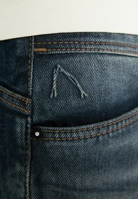 Közelkép kék farmernadrág zsebének, kontrasztos narancsszínű varrással és egy kis fekete szegeccsel az oldalsó varráson.