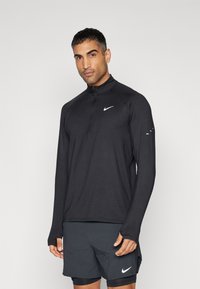 Uomo che indossa una maglietta da corsa nera a maniche lunghe Nike con aperture per i pollici e pantaloni corti neri Nike, in piedi contro uno sfondo chiaro e semplice.