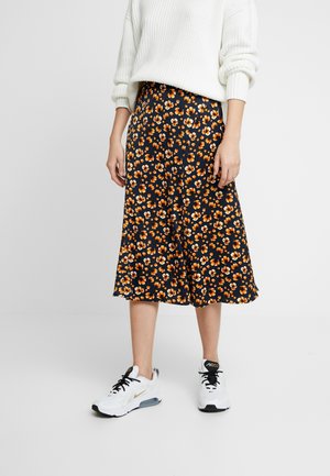 Personne portant un pull blanc, une jupe midi bleu marine à imprimé floral orange et des baskets blanches Nike Air Max sur fond uni.