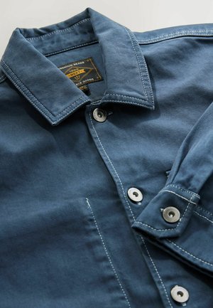 Blue denim jacket with a collared neck, silver buttons, and white stitching details. Features a label on the inside collar.