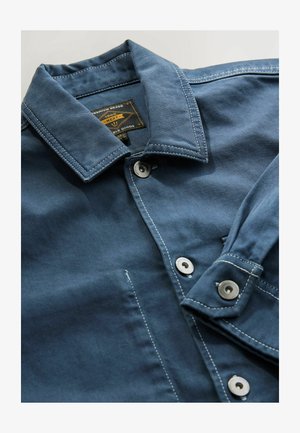 Blaue Jeansjacke mit Kragen, silbernen Knöpfen und weißen Nähdetails. Verfügt über ein Etikett am inneren Kragen.