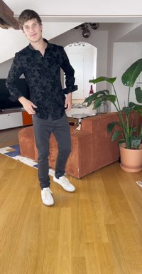Juodas gėlių rašto marškinys, pilkos kelnės ir baltos sportbačiai. Aplinką sudaro tekstūruotas oranžinis sofos ir žalia augalija šviesiame interjere.