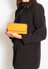 Donna in giacca nera tiene con entrambe le mani una borsa a clutch rettangolare gialla brillante contro uno sfondo neutro.