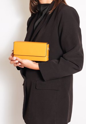 Femme en blazer noir tenant une pochette rectangulaire jaune vif avec les deux mains devant un fond uni.