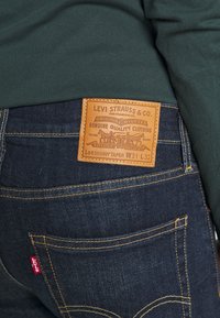 Primer plano del bolsillo trasero de unos jeans Levi's azul oscuro y parche de cuero marrón claro de la marca con los detalles de talla "Lot Skinny Taper W31 L32".