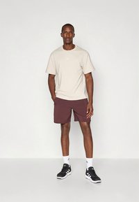 Bēša T-krekls ar Adidas logo, tumši sarkani šorti, melni sporta apavi un baltas zeķes, stāvot pret vienkāršu fonu.