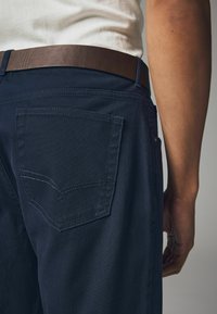 Marineblaue Hosen mit einer strukturierten Oberfläche, die eine Gesäßtasche mit dezentem Stichdetail und einen braunen Ledergürtel um die Taille aufweisen.