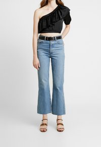 Svart satin crop top med en axel och volanger, högmidjade ljusblå utsvängda jeans och bruna sandaler med leopardmönster och klack.