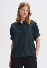 Camicia verde scuro a maniche corte con una texture morbida, chiusura a bottoni e maniche arrotolate, abbinata a pantaloni neri.