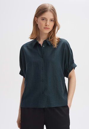 Camicia verde scuro a maniche corte con una texture morbida, chiusura a bottoni e maniche arrotolate, abbinata a pantaloni neri.