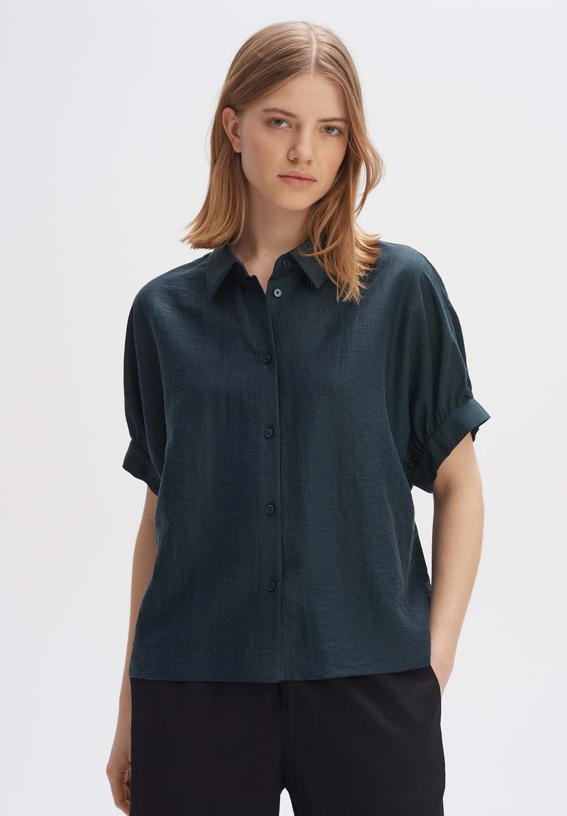 Camicia verde scuro a maniche corte con una texture morbida, chiusura a bottoni e maniche arrotolate, abbinata a pantaloni neri.