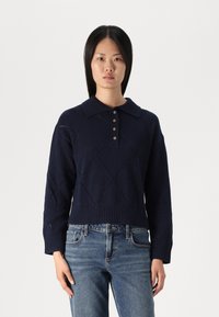 Tamnoplavi pleteni pulover s rebrastim ovratnikom, dugmadi na prednjem dijelu i dijamantskim uzorcima. Skraćena dužina s širokim rukavima.