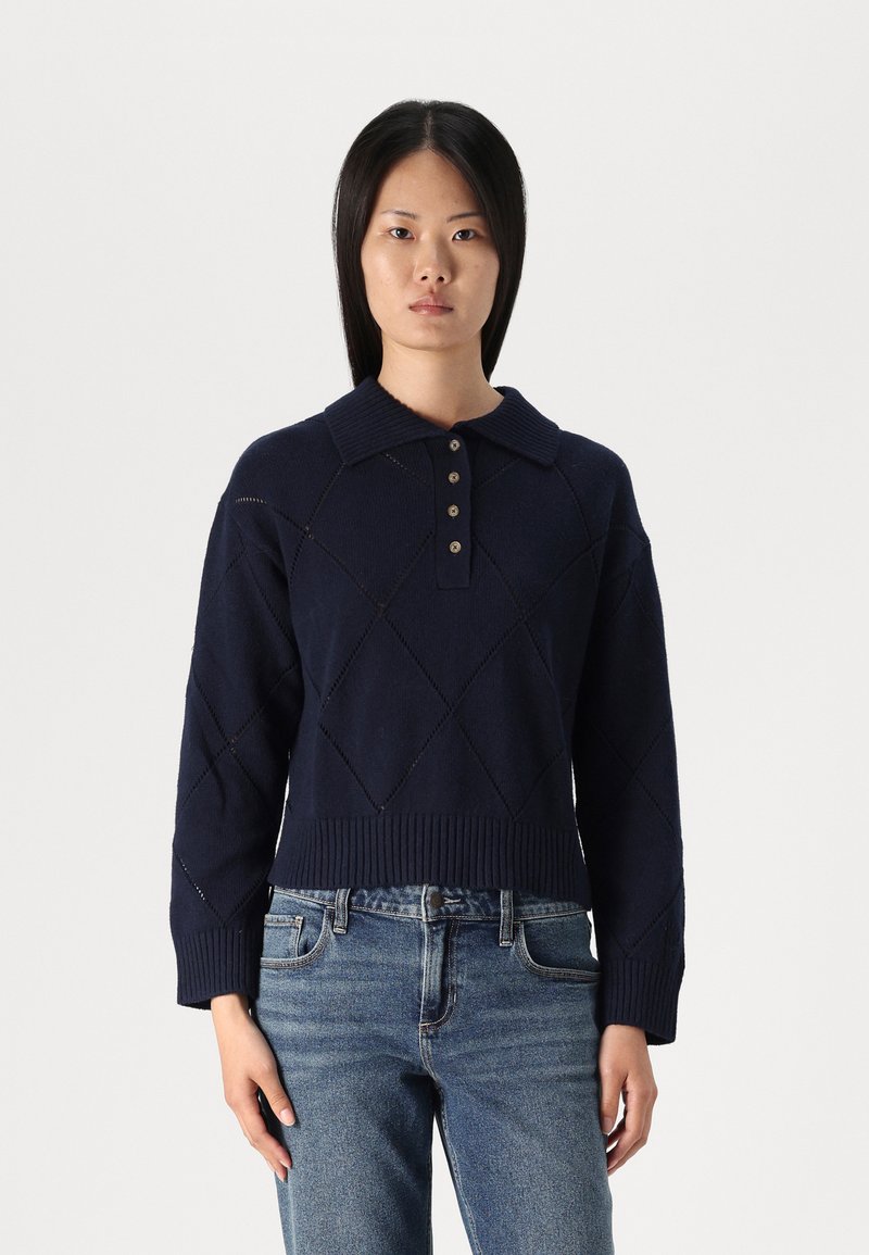 Tamnoplavi pleteni pulover s rebrastim ovratnikom, dugmadi na prednjem dijelu i dijamantskim uzorcima. Skraćena dužina s širokim rukavima.