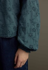 Blusa ricamata turchese con motivi floreali, polsini arricciati e orlo scallop. Abbinata a un tessuto in denim scuro per contrasto.