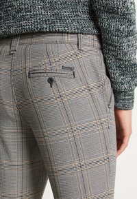 Pantalons à carreaux gris en tissu texturé, avec une poche arrière à boutons et des accents noirs, complétés par un pull en maille verte.