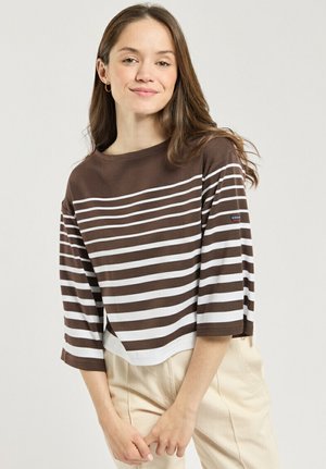Femme aux longs cheveux bruns portant une chemise à rayures marron et blanc à manches trois-quarts et un pantalon beige clair, debout devant un fond uni.