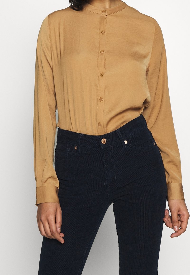 Blouse beige clair à manches longues avec col mandarin et fermeture à boutons sur le devant, accompagnée de pantalons en velours côtelé bleu marine foncé avec une taille à boutons.