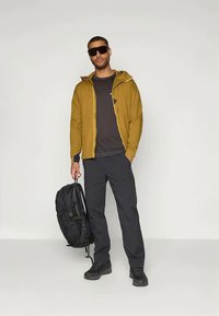 Žuta jakna s kapuljačom, crna majica, tamne hlače i crni ranac. Naočale za sunce i robusne crne cipele upotpunjuju odjevnu kombinaciju.