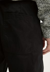 Los pantalones negros cuentan con un gran bolsillo trasero con costuras visibles y una textura suave, combinados con un suéter que muestra un patrón contrastante.