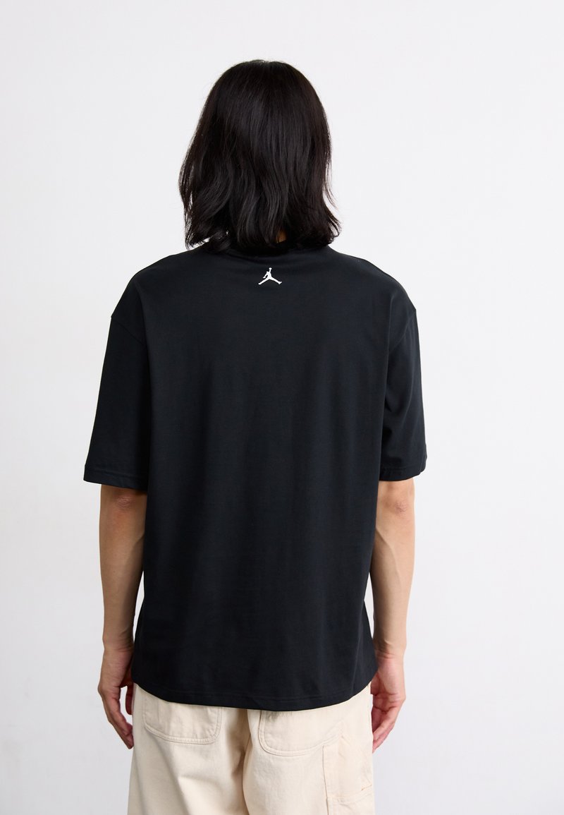 Schwarzes kurzärmliges T-Shirt aus Baumwolle mit lockerer Passform, das ein weißes Jordan-Logo auf dem oberen Rücken aufweist. Weiche Textur.
