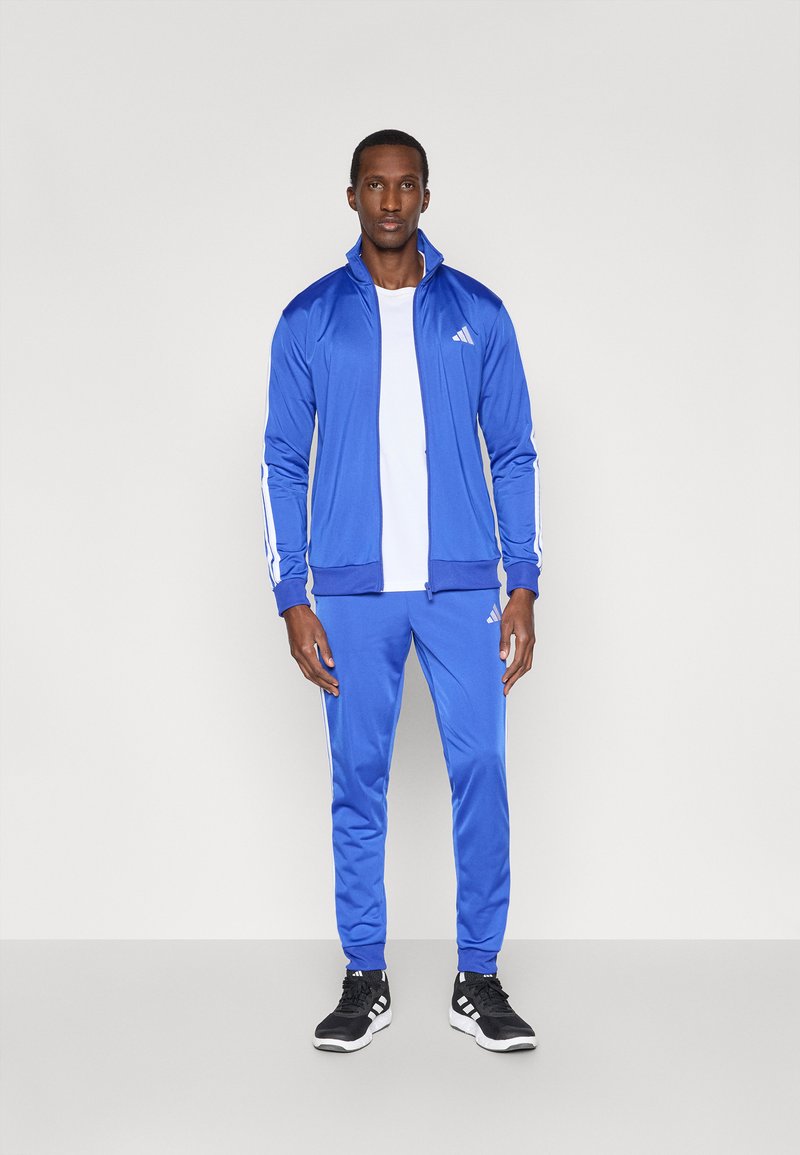 Blauer Sport-Jogginganzug aus glattem Material, mit weißen Seitenstreifen, einer Reißverschlussjacke und schmal zulaufenden Hosen; getragen mit schwarzen Turnschuhen.