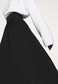 Black pleated skirt with a high waistband, paired with a light-colored long-sleeve top. Features a smooth texture and clean lines.