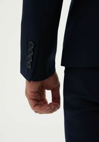 Manche de veste de costume bleu marine montrant un gros plan de quatre boutons noirs et une texture de tissu lisse ; la main repose contre le côté.