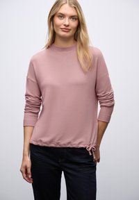 Rosa, weicher Pullover mit hohem Kragen, langen Ärmeln und einem gerafften Kordelzugsaum. Getragen über dunklen Jeans.