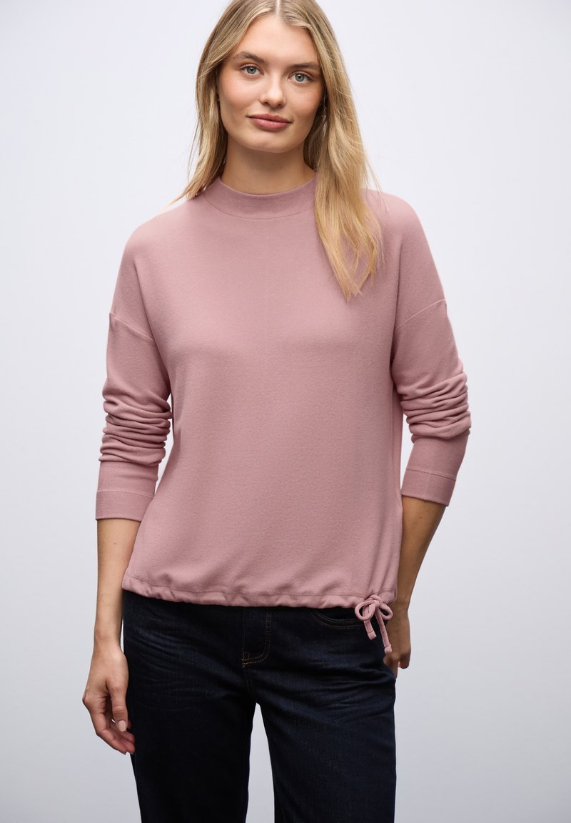 Rosa, weicher Pullover mit hohem Kragen, langen Ärmeln und einem gerafften Kordelzugsaum. Getragen über dunklen Jeans.