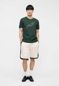 Άνδρας που στέκεται με τα χέρια στις τσέπες, φορώντας σκούρο πράσινο κοντομάνικο πουκάμισο με λογότυπο σε σενάριο, κρεμ σορτς με σκούρες λεπτομέρειες, λευκές κάλτσες και μαύρα παπούτσια.