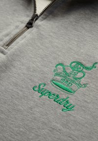 Sudadera gris con media cremallera, que presenta una corona bordada en verde y el texto "Superdry". Textura suave con un diseño casual.