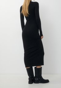 Schwarzes, langärmeliges, figurbetontes Kleid in Midi-Länge. Glatte Textur, taillierte Passform. Kombiniert mit knielangen schwarzen Lederstiefeln.