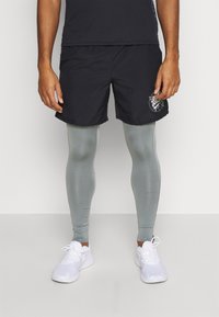 Shorts de sport noirs avec un logo graphique, portés au-dessus de leggings de compression gris clair. La tenue est complétée par des chaussures de sport blanches.