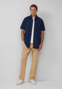 Marineblauw shirt met korte mouwen en twee borstzakken, gecombineerd met beige chino's en witte sneakers, op een neutrale achtergrond.