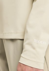 Beige langärmliges Textiloberteil mit strukturiertem Stoff, geradem Saum und einem weißen "TEXADRI"-Logo am Ärmel.
