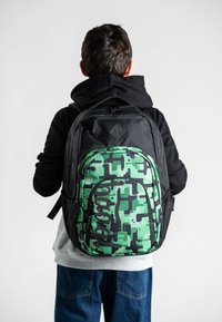 Zaino mimetico verde e nero con un motivo testurizzato, cerniere e spallacci regolabili, indossato da una persona con felpa nera con cappuccio.