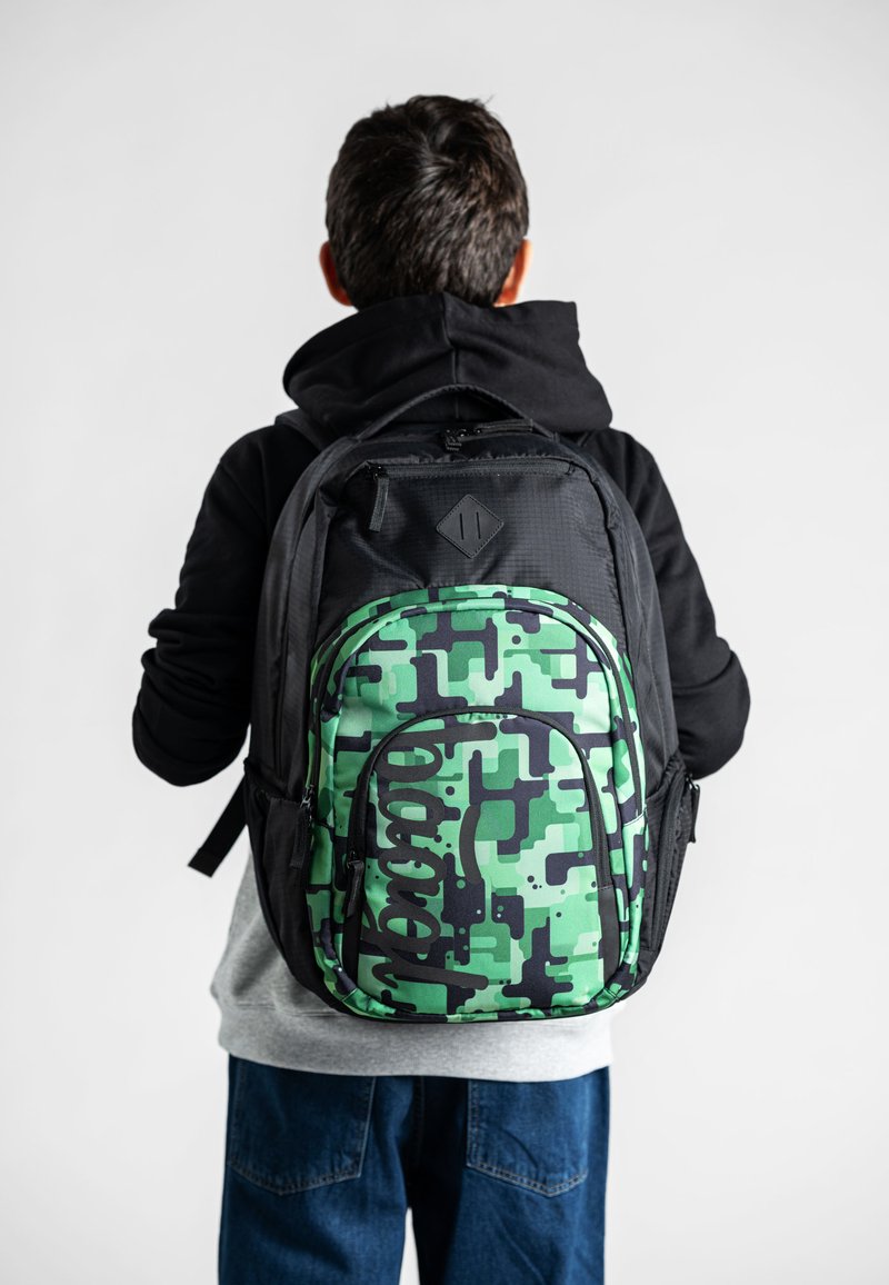Zaino mimetico verde e nero con un motivo testurizzato, cerniere e spallacci regolabili, indossato da una persona con felpa nera con cappuccio.