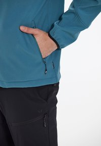 Teal jacka med zipfickor och elastiska manschetter, parat med svarta byxor som har en sidoficka med zippers och subtil logotypdetalj.