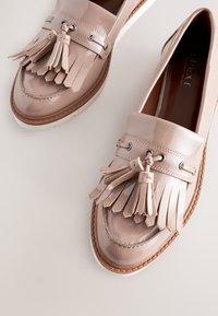 Mocassins en cuir verni beige avec glands à franges, semelle plate blanche et laçage décoratif. Texture lisse et design à bout arrondi.