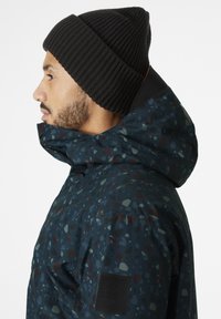 Ciemnoniebieska kurtka z kolorowym plamkowym wzorem, posiadająca wysoki kołnierz, przymocowany kaptur oraz czarną, prążkowaną czapkę beanie.