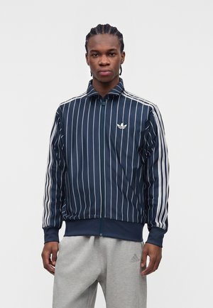 Młody mężczyzna z zaplecionymi włosami, ubrany w granatowo-białą, prążkowaną kurtkę Adidas na zamek oraz jasnoszare spodnie dresowe Adidas, stojący na jednolitym tle.