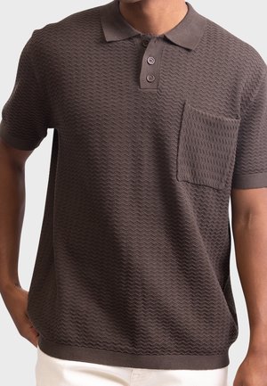Homme portant un polo marron texturé à manches courtes avec deux boutons et une poche poitrine, associé à un pantalon blanc.