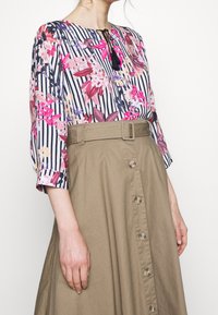 Bluzka w kwiatowy wzór z czarno-białymi paskami, zestawiona z khaki spódnicą z paskiem w talii i zapięciem na guziki z przodu.