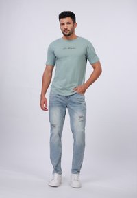 Camiseta de manga corta verde claro con un script negro, combinada con unos vaqueros desgastados azul claro y zapatillas blancas sobre un fondo liso.