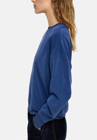 Niebieska bluza z okrągłym dekoltem, długimi rękawami i ściągaczami. Tkanina wydaje się miękka o gładkiej fakturze. Noszona z ciemnymi dżinsami.