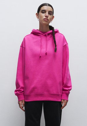 Roze hoodie met een verstelbare koord, ontspannen pasvorm en geribbelde manchet. Draagt met donkere broeken, wat de gladde, zachte stofstructuur benadrukt.